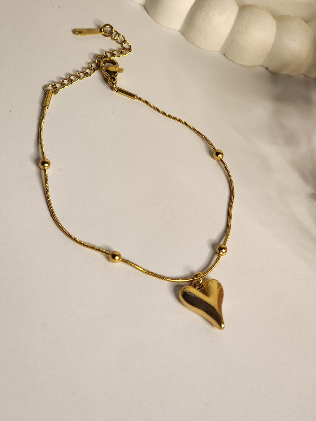 Gold puffy heart charm ball station snake chain bracelet laid flat on white surface