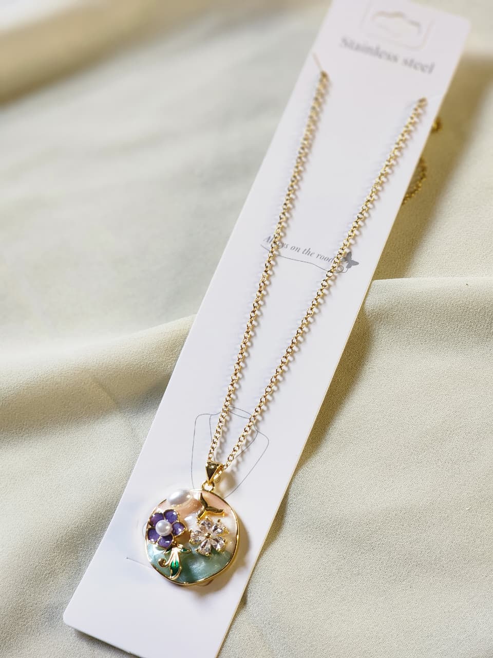 Gold floral pendant necklace with mother-of-pearl shell background and purple flower crystal accents on packaging card