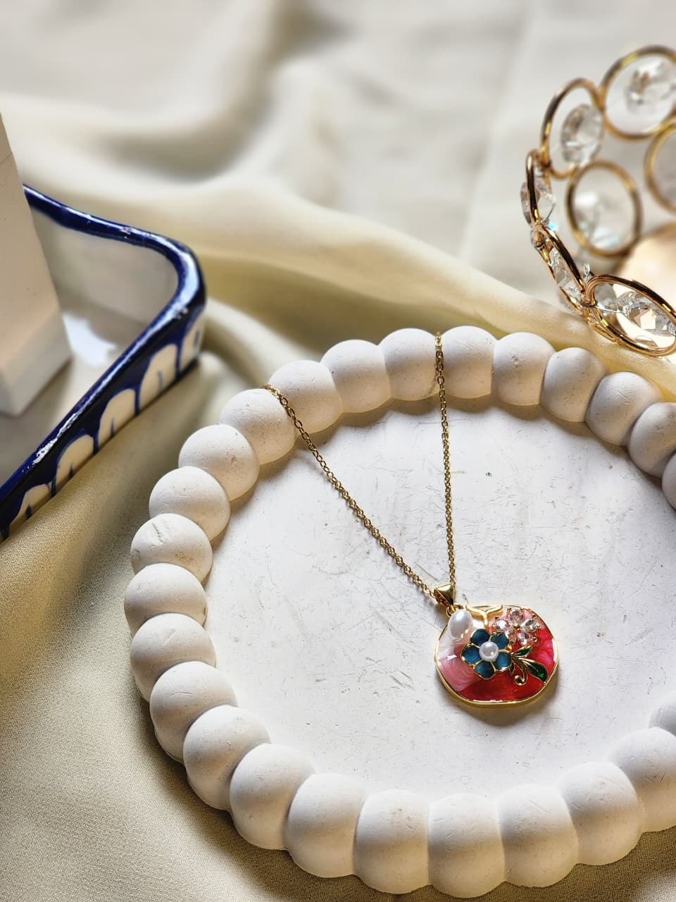 Gold floral enamel pendant necklace with red background and blue flower charm on white display tray