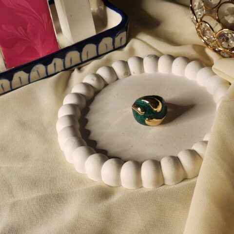 A high-angle shot of a bold dome-shaped statement ring featuring a deep emerald green malachite texture with swirling polished gold inlays, displayed on a white beaded decorative tray.