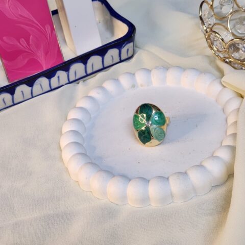 A high-angle shot of a polished gold oval statement ring featuring a four-leaf clover pattern in marbled shades of emerald and seafoam green, centered with a sparkling crystal.