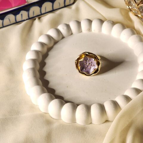 A close-up shot of a single-bloom statement ring featuring lilac, set on a premium gold finish base and displayed on a white decorative tray.