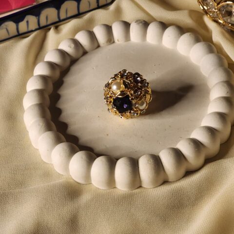 A luxurious dome-shaped statement ring featuring a cluster of deep purple amethyst-style stones, white pearls, and shimmering crystals set in an ornate gold floral frame.