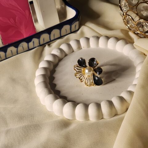 A striking gold statement ring featuring three black enamel petals and a shimmering white pearl center, accented by a decorative gilded scallop fan.