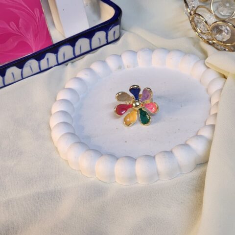 A colorful gold-finish statement ring featuring a multi-color rainbow petal design on a white decorative tray.