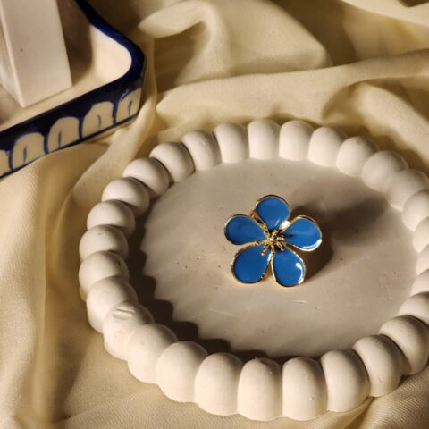 A close-up shot of a gold-finish statement ring featuring five vibrant blue petals on a white beaded decorative tray.