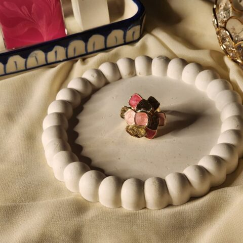 A top-down shot of a gold-finish statement ring featuring a cluster of textured gold petals and smooth pink enamel petals on a white decorative tray.