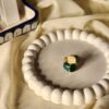 A high-angle shot of a thick gold-finish statement ring featuring two large, rounded square stones in cream and deep malachite green, displayed on a white beaded decorative tray.