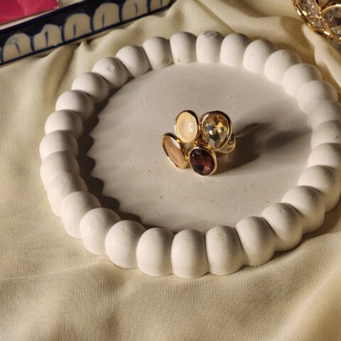 A high-angle shot of a gold-finish statement ring featuring a cluster of four oval stones in cream, amber, and deep plum, displayed on a white decorative tray.