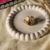 A high-angle shot of a gold-finish statement ring featuring a cluster of four oval stones in cream, amber, and deep plum, displayed on a white decorative tray.